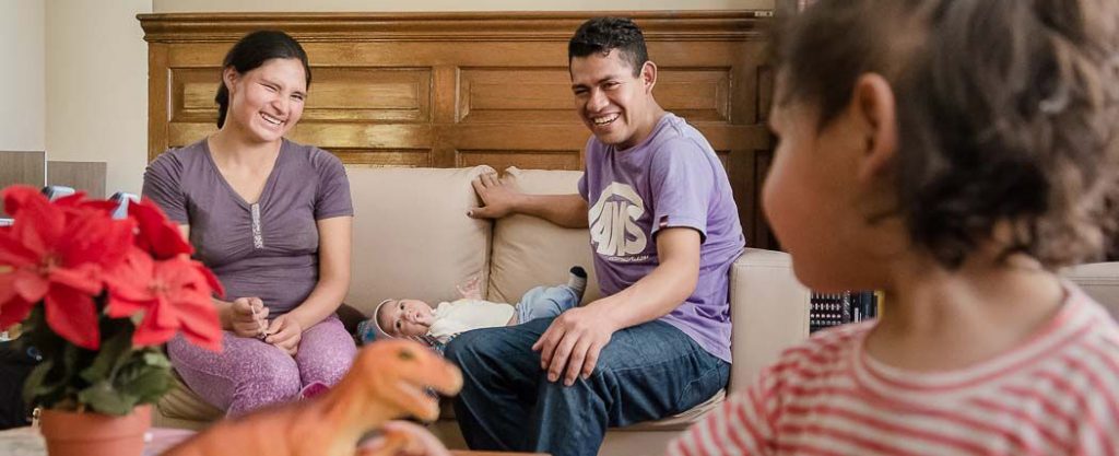 Padre y madre sentados con su bebe en acostado en medio de ellos. Ambos miran a una niñita, frente a ellos, quien los mira también.